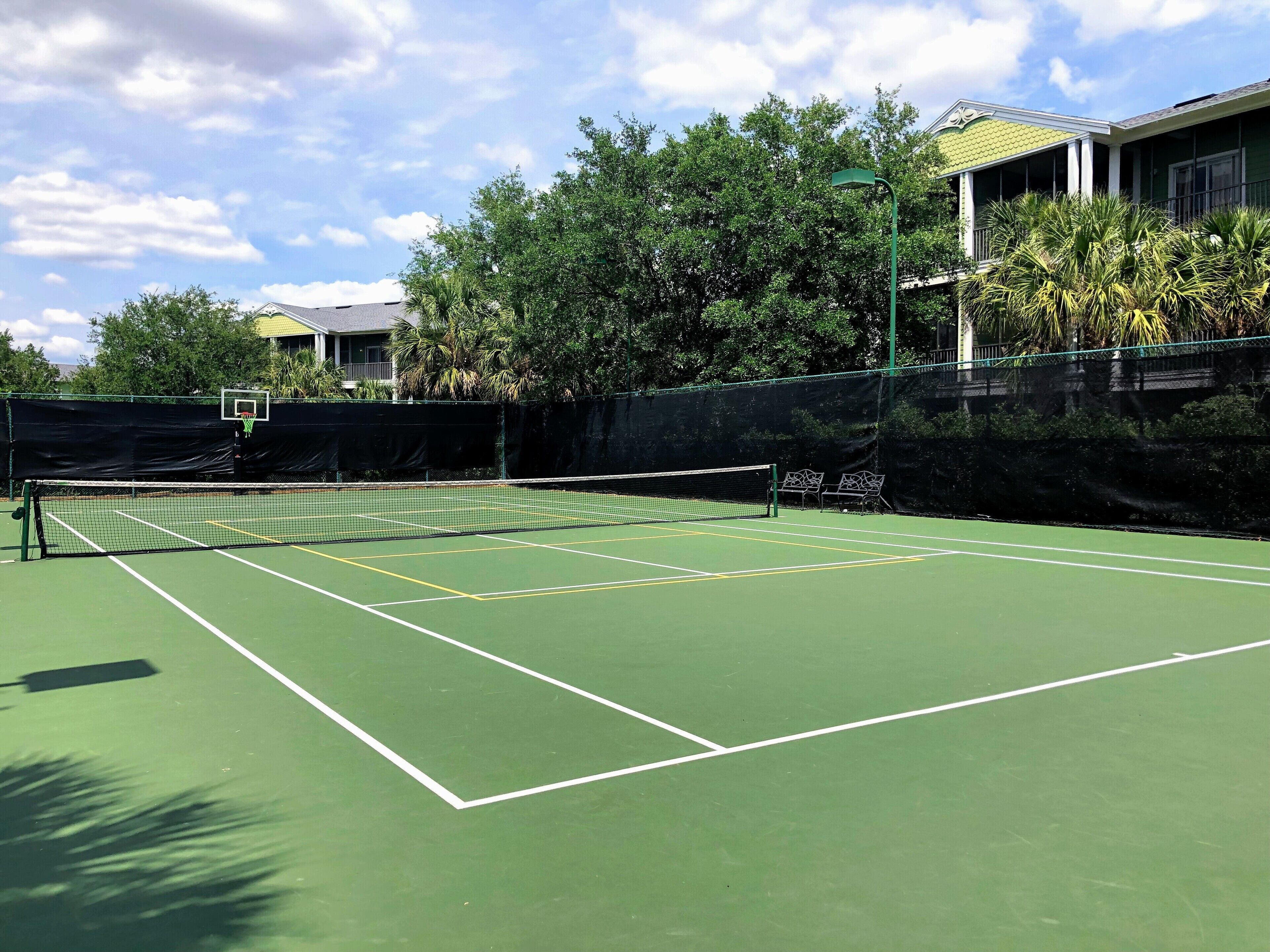 A tennis court with a net, surrounded by trees and a building with a green roof.