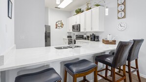 Eine moderne Küche mit weißen Schränken, Edelstahlgeräten und einer weißen Arbeitsplatte. Zwei Barhocker mit Ledersitzen und Holzbeinen sind vorhanden.