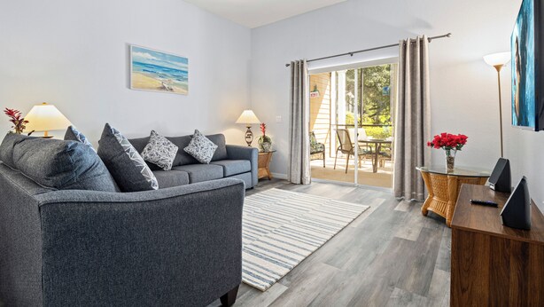 Ein modernes Wohnzimmer mit einem grauen Sofa, zwei Beistelltischen mit Lampen, einem großen Fenster mit Vorhängen und Blick ins Freie.