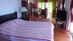 A hotel room with a large bed, a TV, a desk, and a chair. There is a window with curtains and a plant on the desk. - Family Suite Sea View (Mahe)