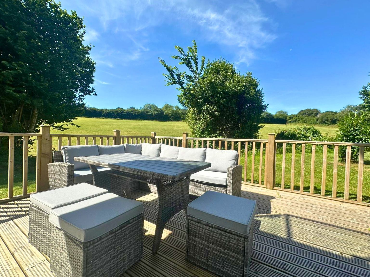 Une terrasse extérieure avec un ensemble de meubles de jardin en osier (canapé et table), entourée d’une plateforme en bois et d’une clôture en bois.