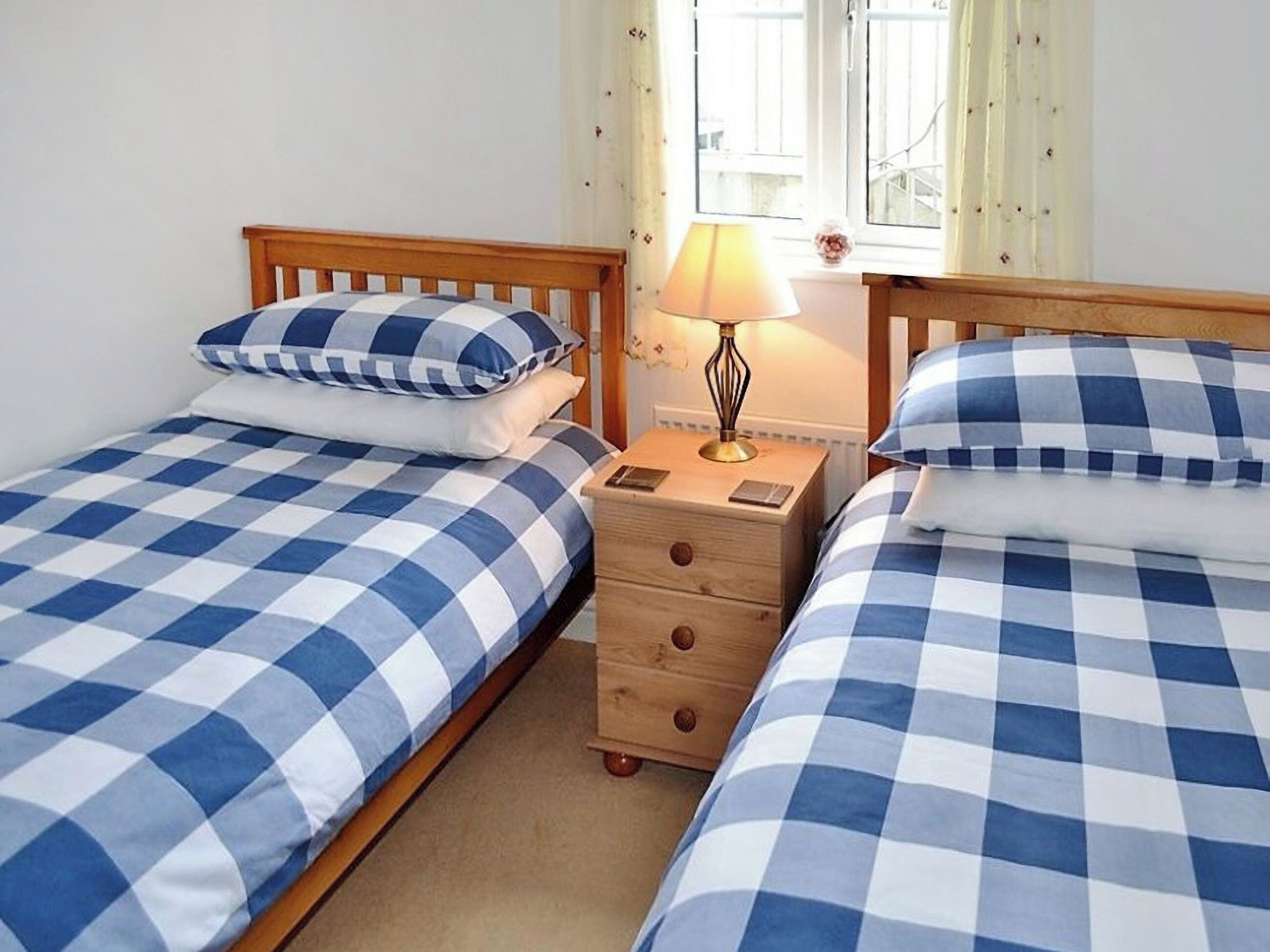 Une chambre à coucher avec deux lits, chacun recouvert d’une couette à carreaux bleus et blancs, une table de chevet avec une lampe et une fenêtre avec des rideaux.