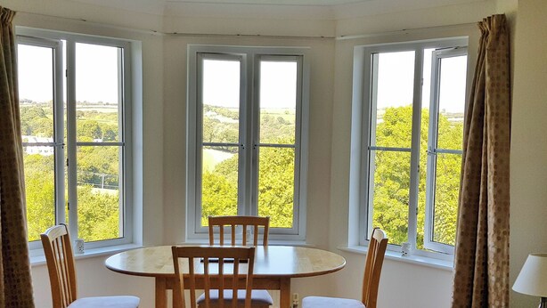 A dining area with a round table and four wooden chairs, large windows with curtains, and a view of greenery outside. - Meadowside Apartment (Fowey)
