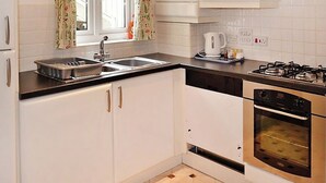 A kitchen with white cabinets, a gas stove, a sink, and a window with floral curtains. - Meadowside Apartment (Fowey)