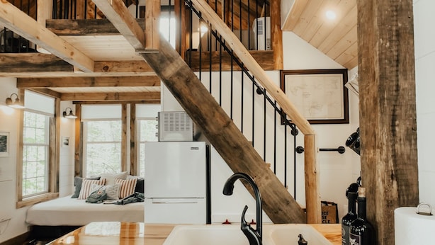 Un interior rústico con vigas de madera a la vista, una escalera de madera, una zona de comedor con mesa y sillas, y una cocina con un fregadero y encimera.