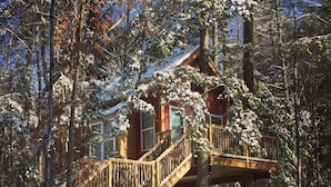 Una cabaña con terraza de madera y escaleras que conducen a un balcón en un segundo piso, rodeada de árboles cubiertos de nieve.