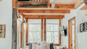Interior acogedor de una cabaña con habitación en el entrepiso, vigas de madera, un ventanal grande, un sofá y una mesita de centro.