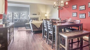 Un área de comedor con pared roja, suelo de madera y una araña. Se observa una chimenea y un sofá al fondo.