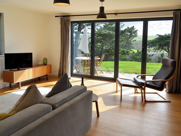 A modern living room with a sofa, a chair, a TV, and a large window offering a view of a garden.