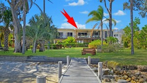 Un resort con una terraza de madera que conduce a un edificio con varios balcones, rodeado de palmeras y un césped bien cuidado.