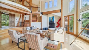 A spacious living area with a high ceiling, wooden beams, a fireplace, and a large window.