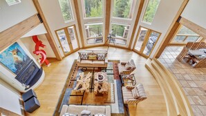 A spacious loft-style living area with a large central seating area, multiple windows offering views of a forested area, and a dining area with wooden furniture.