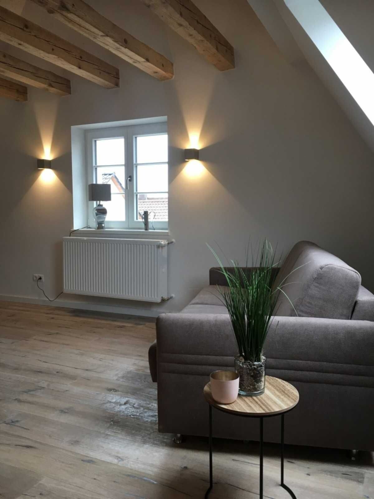 A loft-style room with exposed wooden beams, a grey sofa, a small round table with a plant, and two wall-mounted lights.