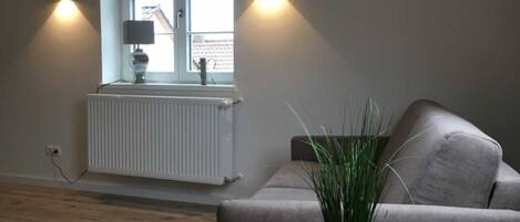 A loft-style room with exposed wooden beams, a grey sofa, a small round table with a plant, and two wall-mounted lights.