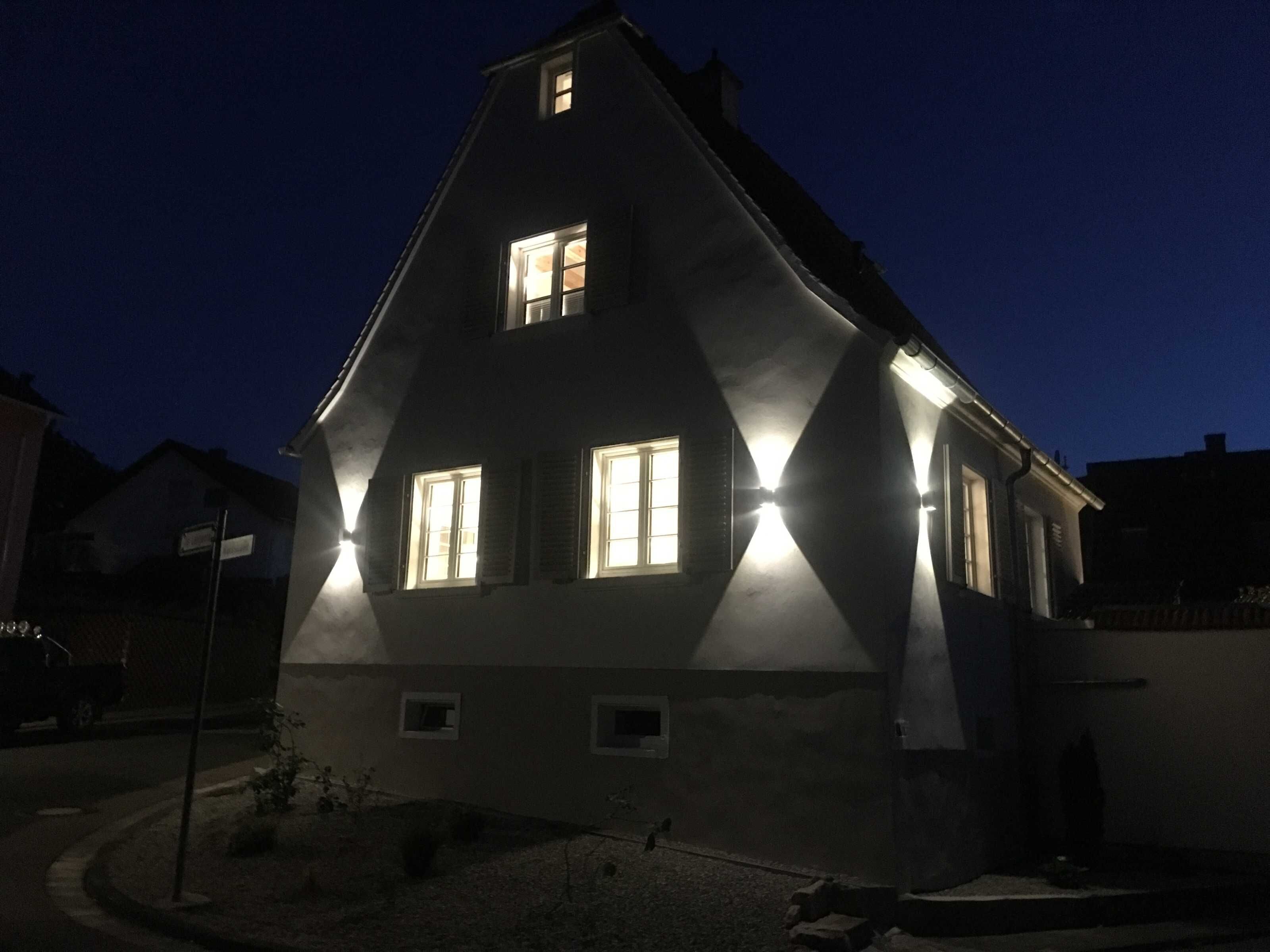 Une maison de couleur sombre, dotée de deux fenêtres visibles, l’une à l’étage supérieur et l’autre au rez-de-chaussée, éclairées par un éclairage extérieur.