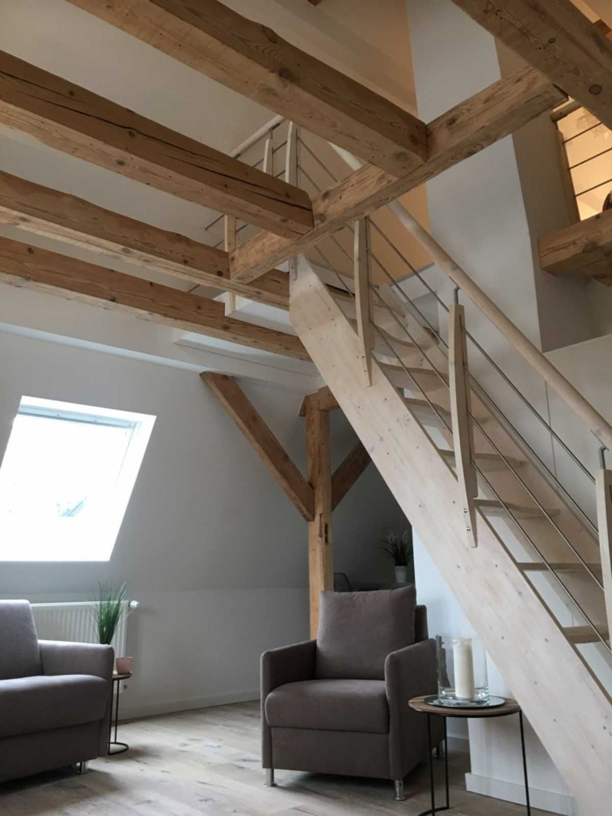 Une pièce de style loft avec des poutres en bois apparentes, un escalier, un fauteuil gris, une petite table avec une bougie et une fenêtre avec des stores.