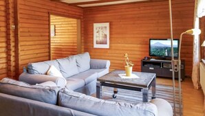 Interior de cabana de tronco com sofá seccional, mesa pequena, televisão e luminária.
