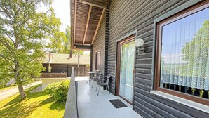 Uma cabana de madeira com varanda, uma mesa com cadeiras e uma janela grande com cortinas de renda.