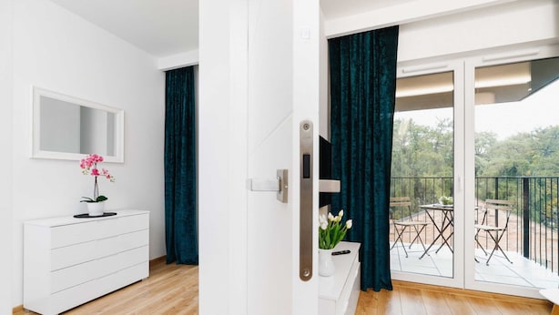 Ein modernes Zimmer mit einer weißen Kommode, einem Spiegel und einer Topfpflanze. Eine Glastür führt auf einen Balkon mit Tisch und Stühlen.
