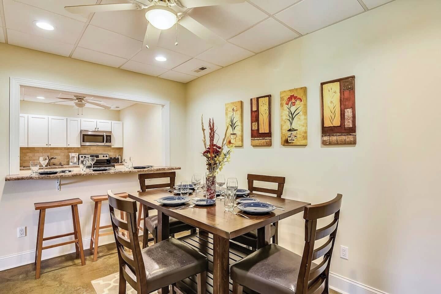 Un comedor con una mesa de madera dispuesta para cuatro personas, rodeada de sillas. Se ve una cocina con armarios blancos y un microondas al fondo.