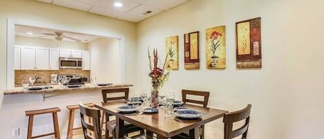 Un comedor con una mesa de madera dispuesta para cuatro personas, rodeada de sillas. Se ve una cocina con armarios blancos y un microondas al fondo.