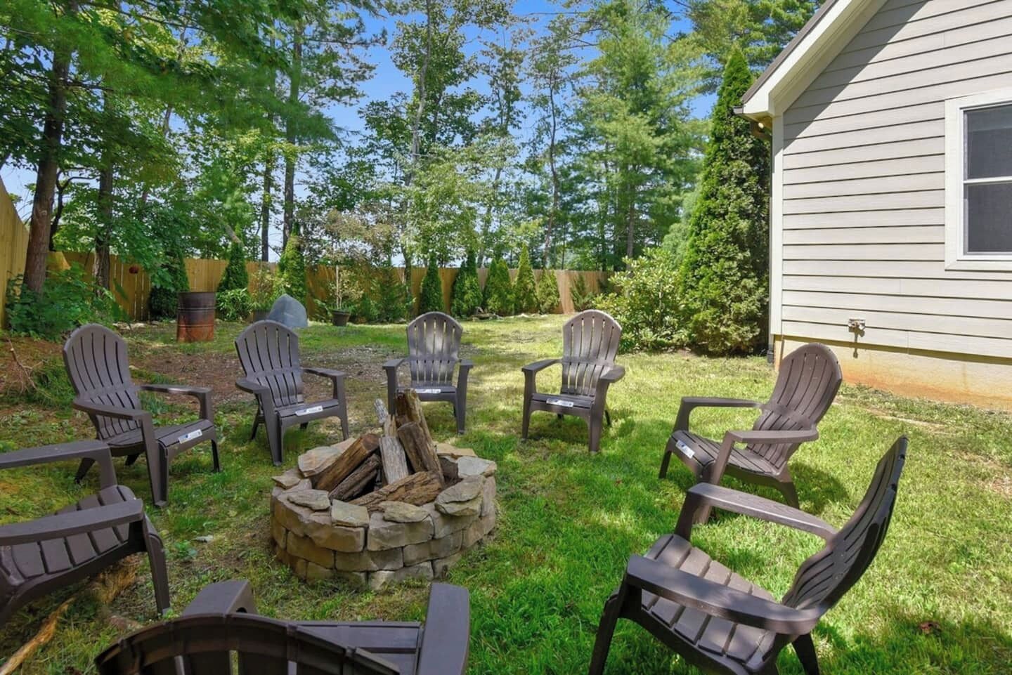 Un patio trasero con un brasero rodeado de sillas Adirondack, una terraza de madera y una casa al fondo.