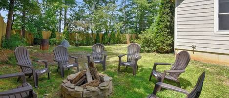 Un patio trasero con un brasero rodeado de sillas Adirondack, una terraza de madera y una casa al fondo.