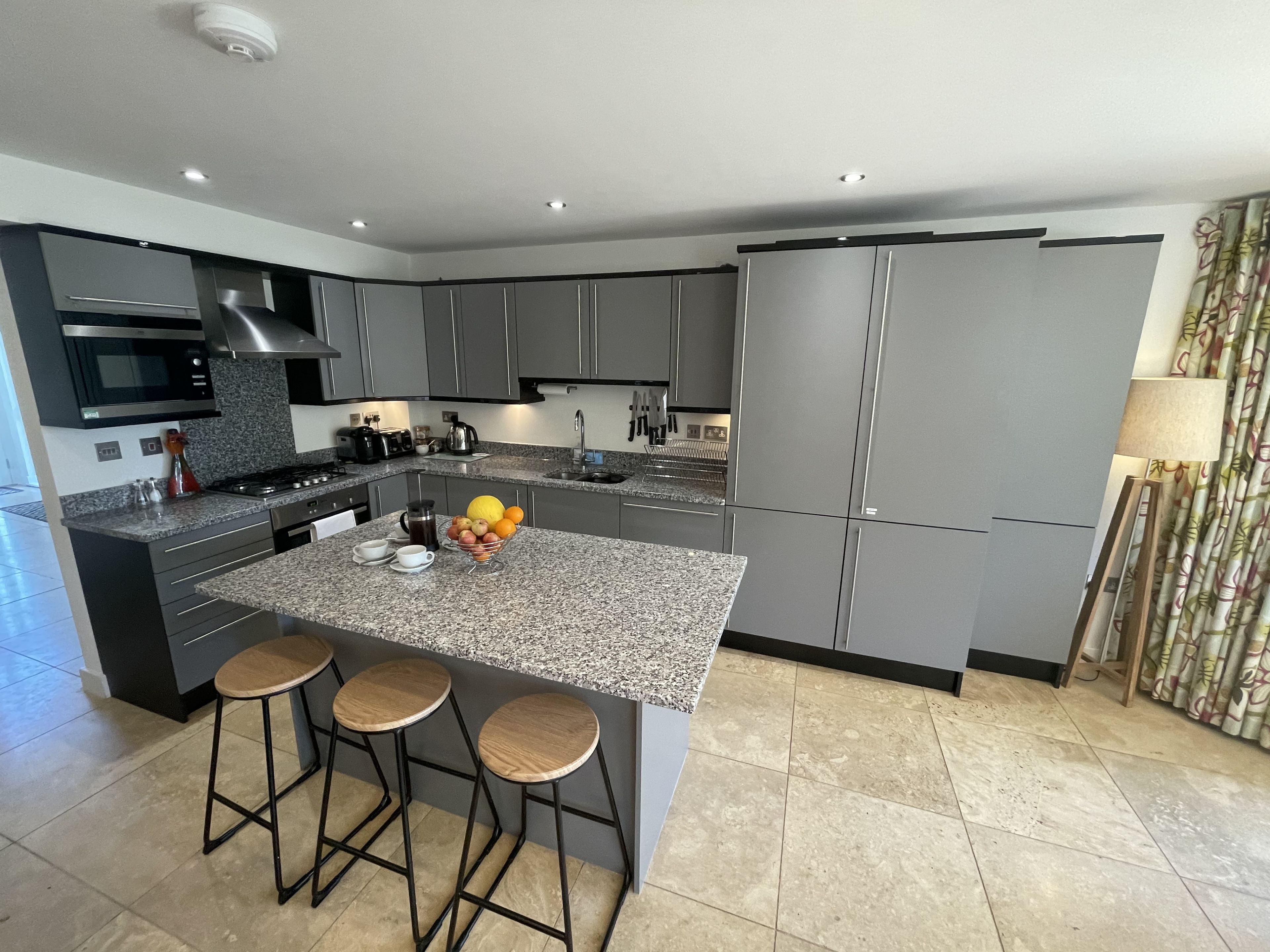 A modern kitchen with grey cabinetry, a granite island with stools, and stainless steel appliances.