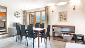 A dining area with a table and chairs, a clock, a window with curtains, and a wall-mounted shelf with baskets. - 800m des Remontées ! Appart Rustique + Sauna (Gresse en Vercors)