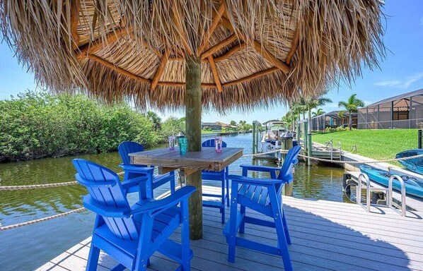 A wooden deck with a thatched roof table and blue chairs overlooking a water body, with buildings and greenery in the background. - Bellissimo - luxury home pool and Jacuzzi, Kayaks (Cape Coral)
