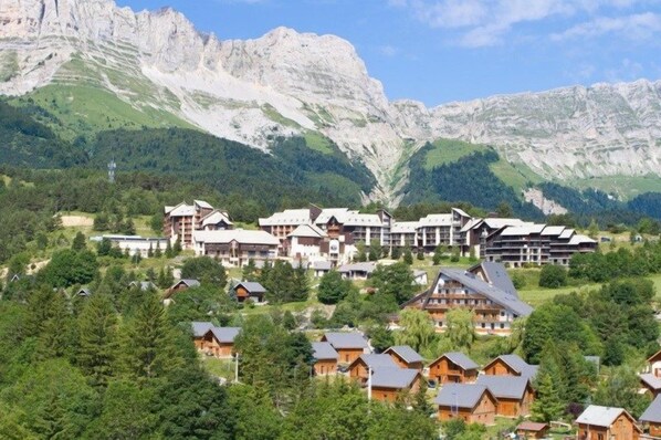 Une station de montagne dotée de chalets et d’un grand bâtiment à multiples balcons, le tout entouré d’une végétation luxuriante et surplombé par d’imposants sommets.