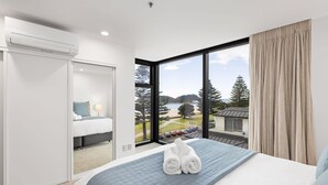 A modern hotel room with a large bed, a view of a beach and trees, and a separate bathroom area. - Oceanside Bliss Absolute Beachfront Stunning Views (Mount Maunganui)