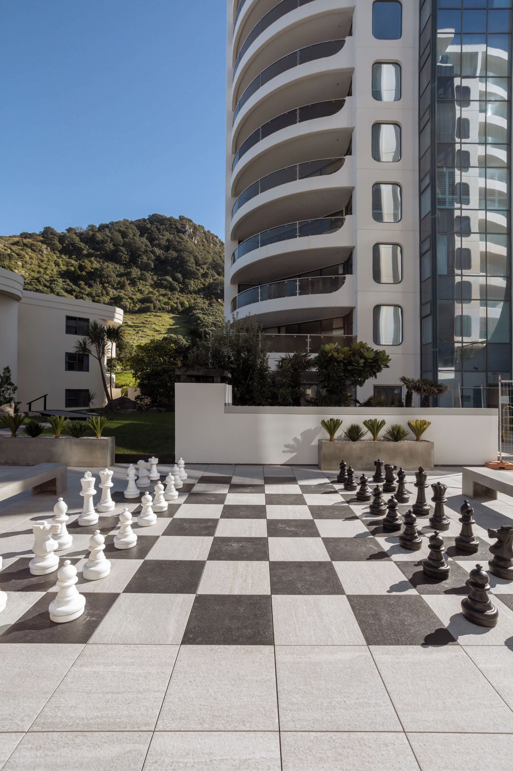 Un échiquier d’extérieur à carreaux avec des pièces d’échecs, un bâtiment moderne doté de balcons, et une colline en arrière-plan.