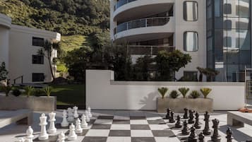 Un échiquier d’extérieur à carreaux avec des pièces d’échecs, un bâtiment moderne doté de balcons, et une colline en arrière-plan.