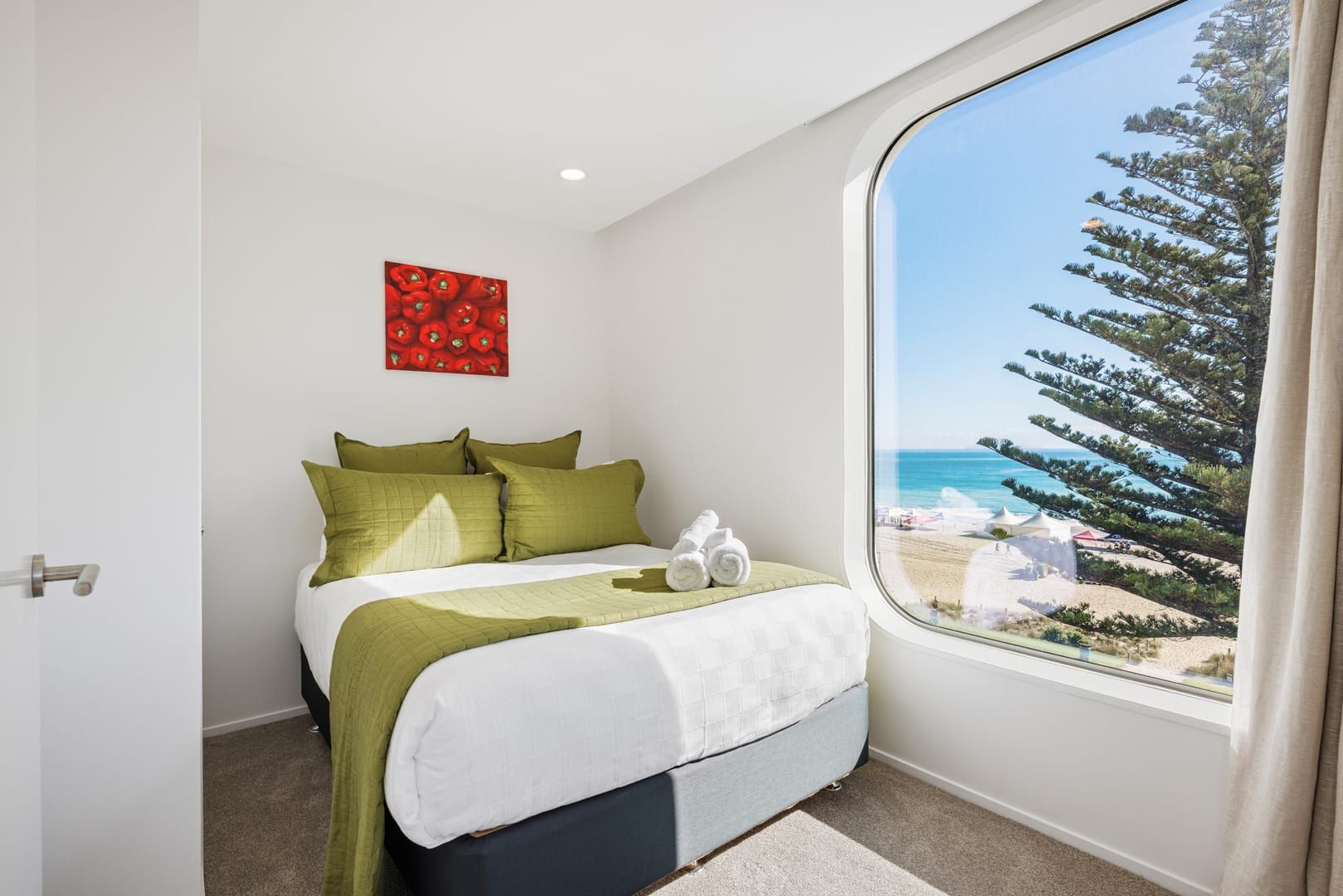 Une chambre à coucher avec un lit, des oreillers verts, un couvre-lit blanc, une œuvre d’art rouge et, par la fenêtre, une vue sur une plage et des arbres.