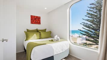 Une chambre à coucher avec un lit, des oreillers verts, un couvre-lit blanc, une œuvre d’art rouge et, par la fenêtre, une vue sur une plage et des arbres.