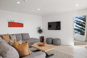 A modern living room with a grey sofa, a coffee table, and a TV mounted on the wall. - Breakwater - Views, Pool, Sauna and Gym (Mount Maunganui)