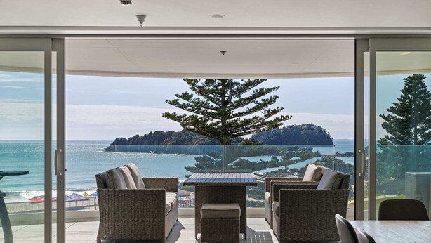 A balcony with wicker furniture, a glass door, and a view of the sea and a distant island. - Oceanside Bliss Absolute Beachfront Stunning Views (Mount Maunganui)