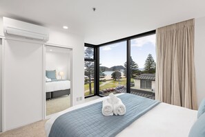 Una camera d'albergo moderna con un'ampia finestra, un letto con copriletto blu e vista sulla spiaggia e sugli alberi.