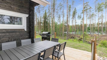 A wooden deck with a table and chairs, a grill, and a view of a forest and lake.