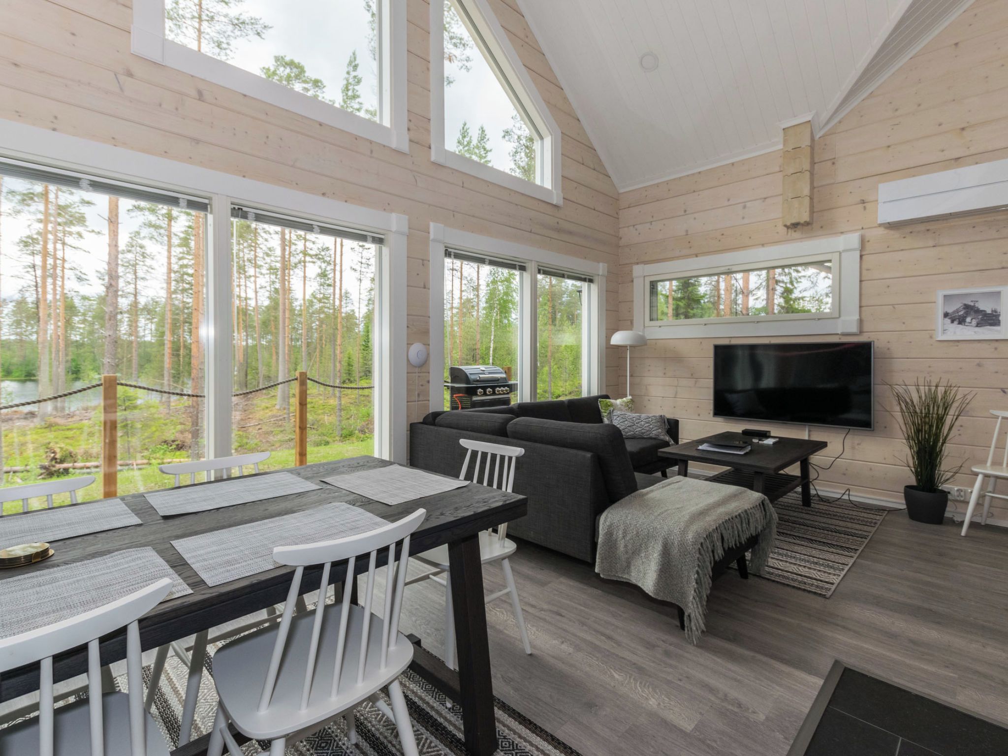 A modern cabin interior with a dining area, sofa, TV, and large windows overlooking a forest.