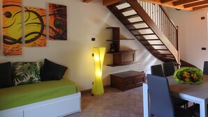 Une pièce avec un escalier en bois, un canapé avec des coussins verts, une table à manger avec des chaises et des œuvres d’art abstraites accrochées au mur.