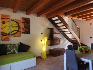 Um quarto com escada de madeira, sofá com almofadas verdes, mesa de jantar com cadeiras e arte abstrata na parede.