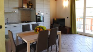 Une cuisine moderne avec des meubles blancs, une cuisinière et un four. On y trouve une table à manger avec des chaises et un bouquet de fleurs. Un téléviseur est posé sur un meuble près d’une fenêtre ornée d’un rideau.