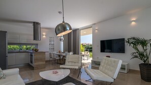 A modern living room with a white sofa, a coffee table, and a dining area with a table and chairs. There is a kitchen with a stove, sink, and cabinets. A large plant is in the corner, and a TV is mounted on the wall. - Beautiful villa with private pool, 3 bedrooms, 3 bathrooms, near the seaside! (Fazana)