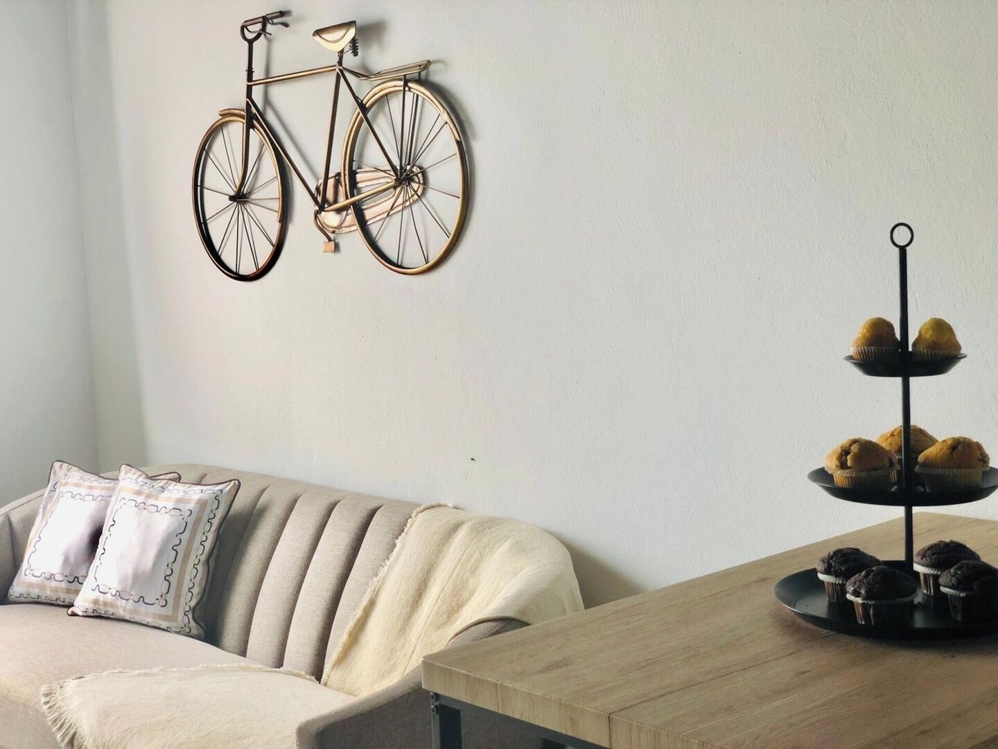Une pièce avec un canapé beige, une table basse en bois, un vélo fixé au mur et un présentoir à étages contenant des muffins.