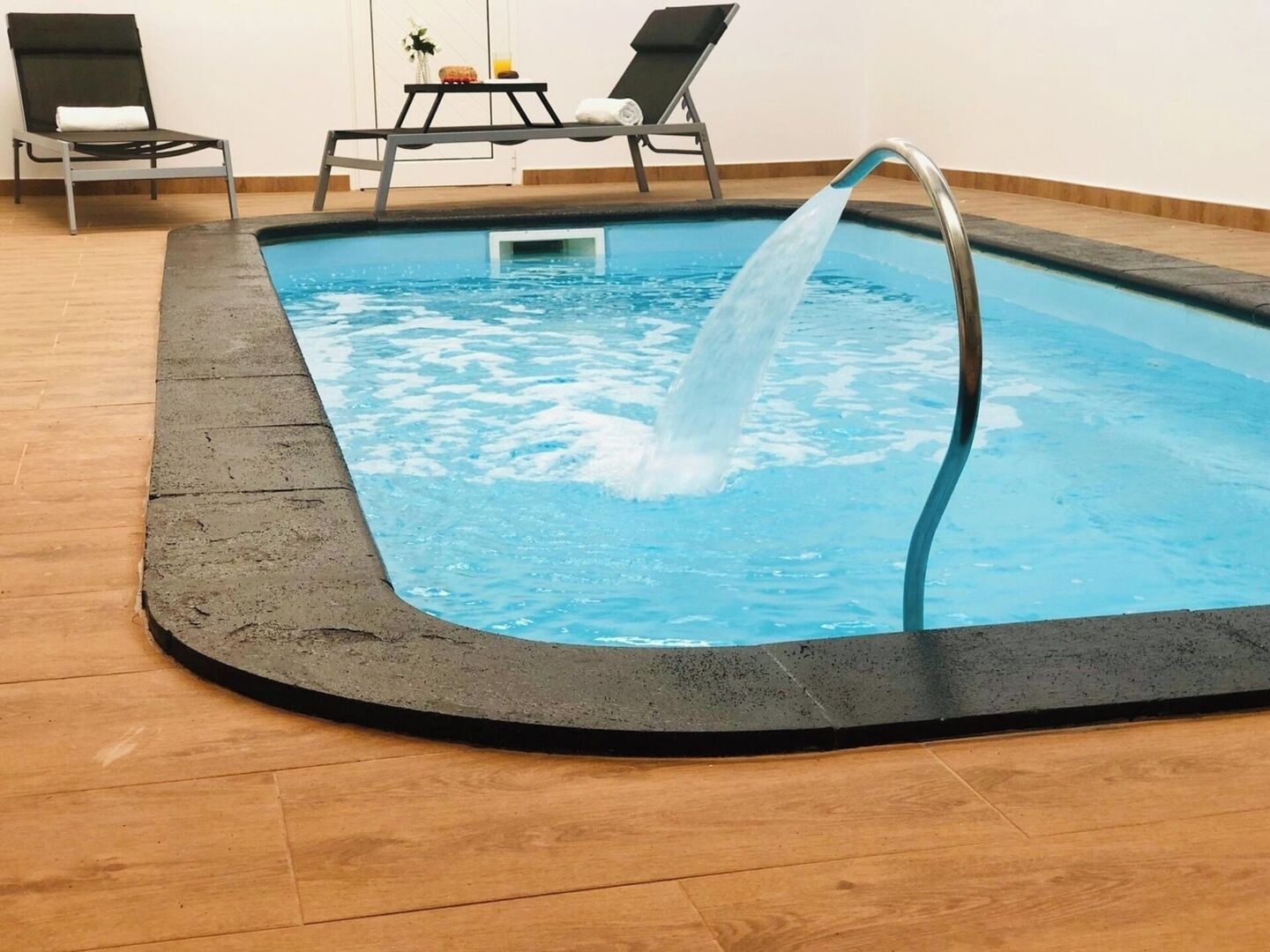 Piscina interna con fontana, due sdraio e un tavolino con una pianta e un bicchiere.
