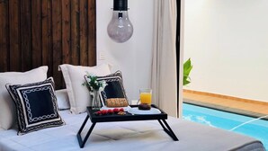 A bedroom with a bed, pillows, a bedside table with a glass of orange juice, and a view of a swimming pool. - Canaryislandshost l Nazaret Suites (Nazaret)