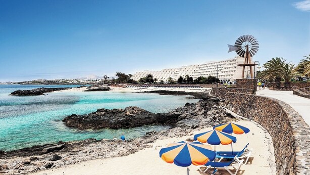 A coastal beach with clear turquoise waters, a windmill, and a row of beach umbrellas. - Canaryislandshost l Nazaret Suites (Nazaret)
