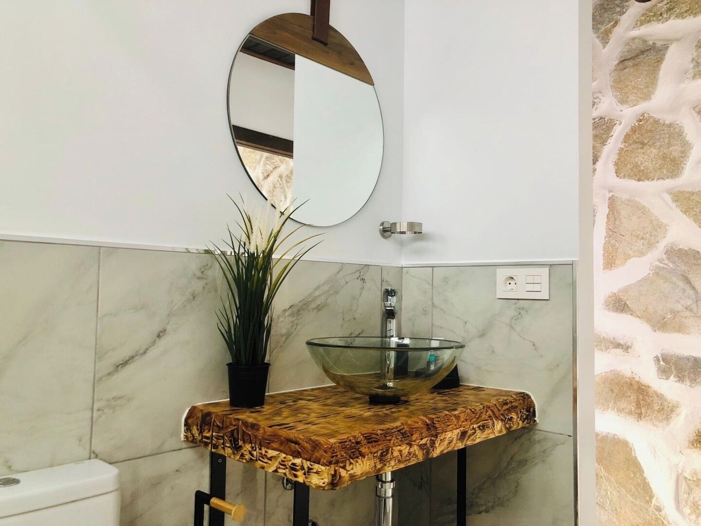 A bathroom with a marble wall, a round mirror, a glass sink, and a potted plant.